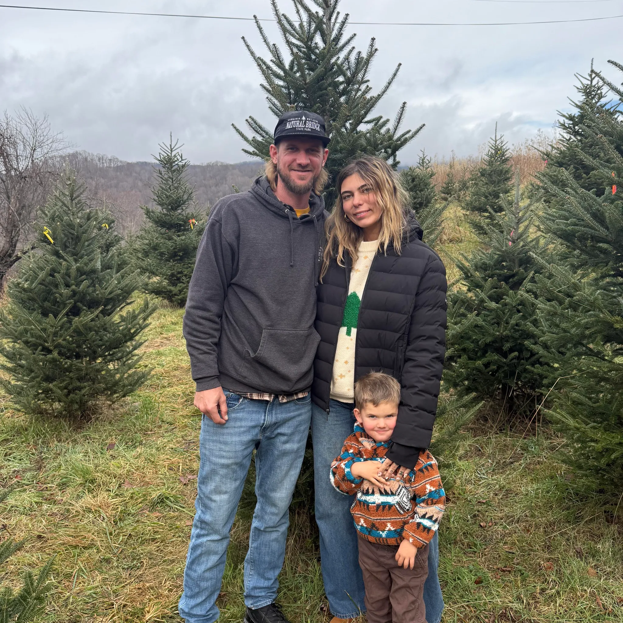 Josh, Sage, and their son standing in a Western North Carolina field with the Blue Ridge in the distance.